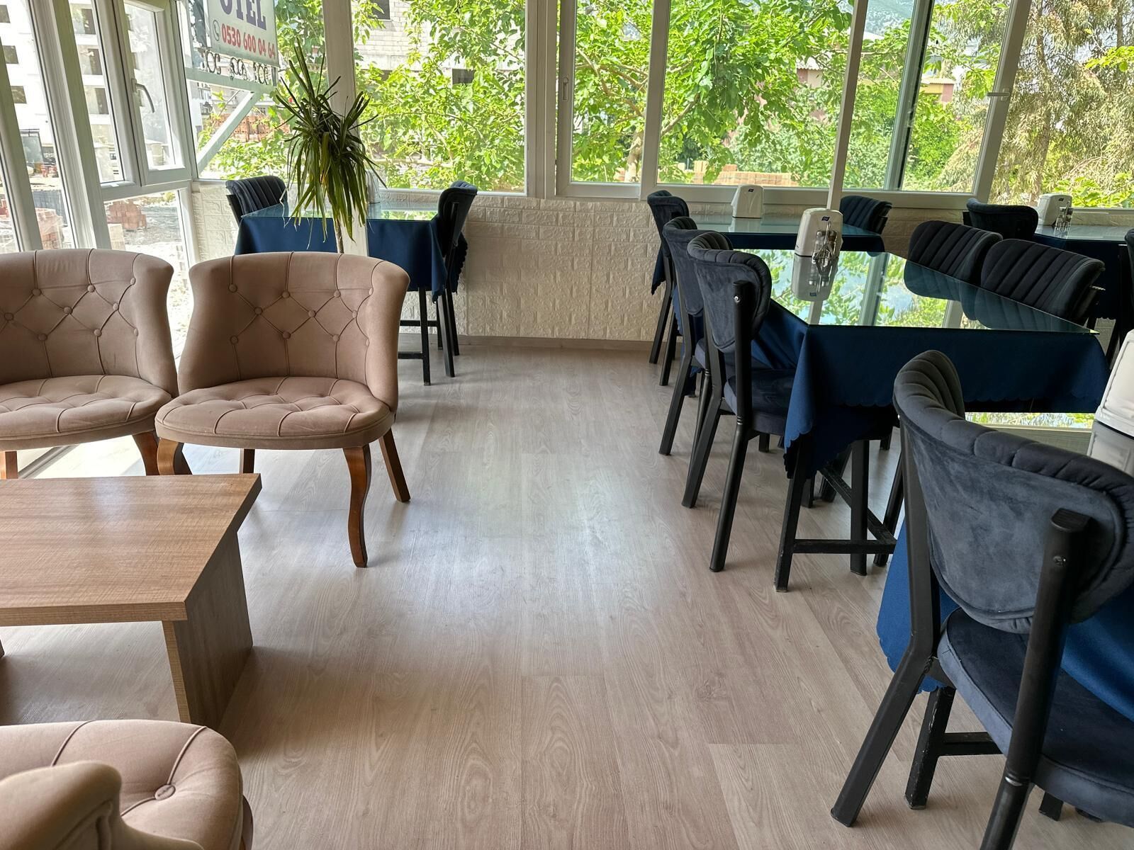 A modern interior with a long table and several chairs, large windows with a view of greenery, and a potted plant.