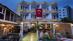 Un hotel con una bandera roja, mesas al aire libre y una entrada bien iluminada.
