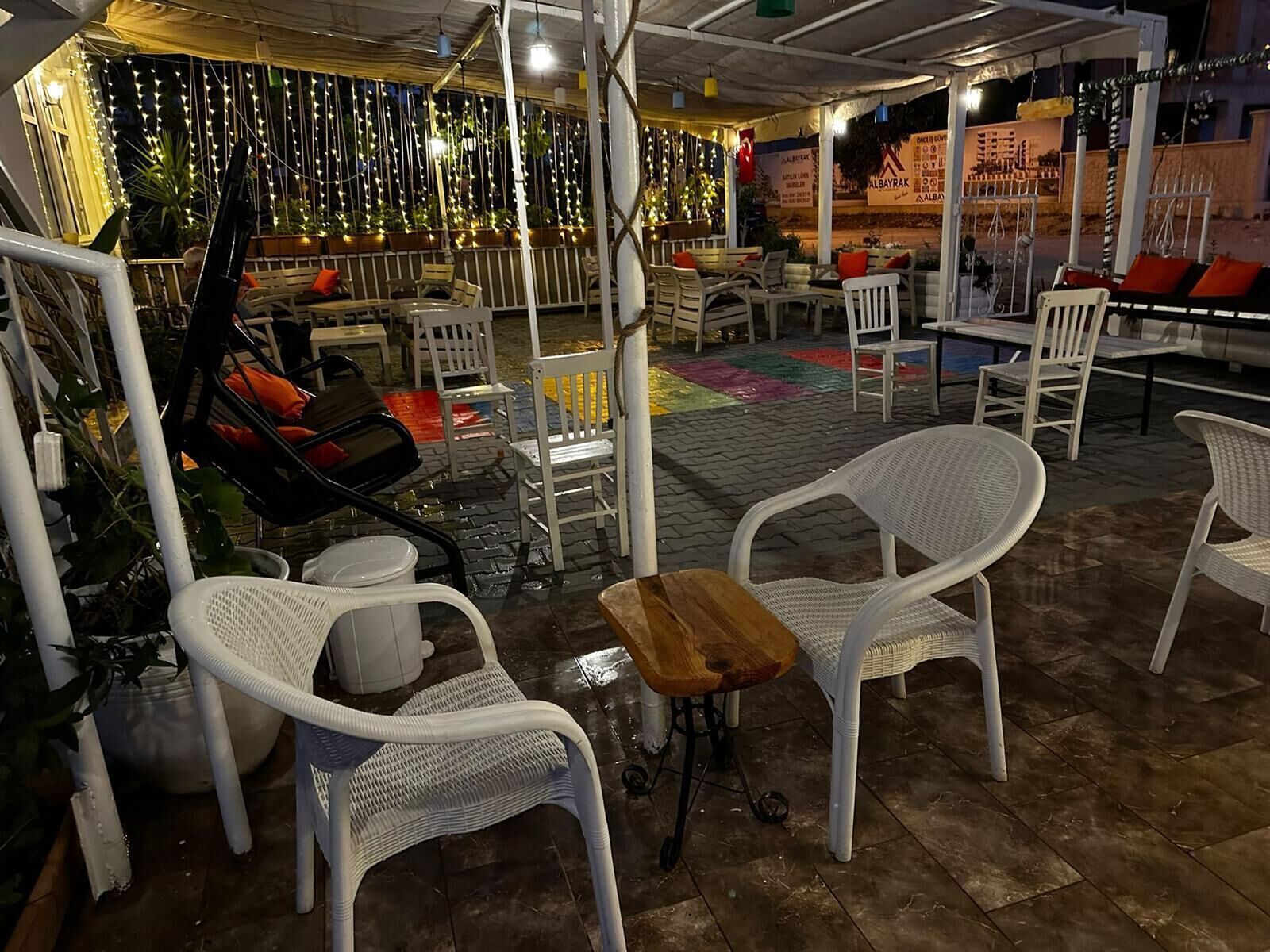 An outdoor seating area with white wicker chairs and tables, surrounded by potted plants and string lights.
