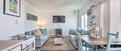 Un salon moderne avec un canapé, une table basse et une table à manger. Un téléviseur est fixé au mur, il y a une petite table d’appoint et une fenêtre avec des rideaux.