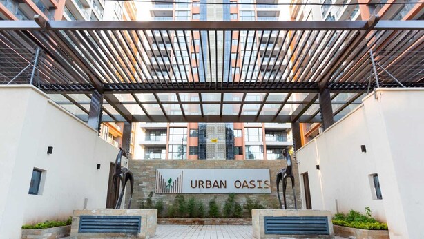 A modern urban building with a covered entrance, a sign reading "URBAN OASIS," and a view of surrounding high-rise buildings.
