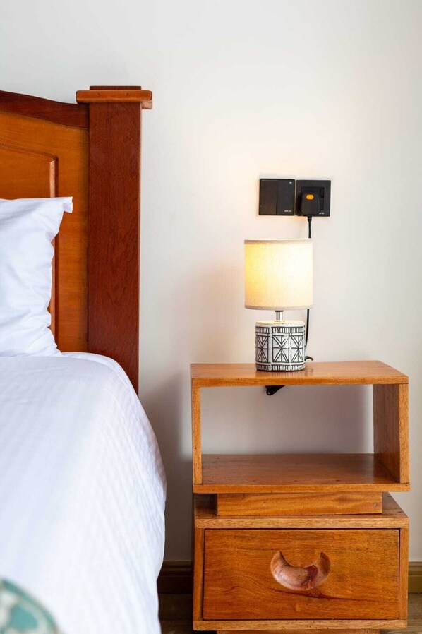 Une table de chevet en bois, avec une lampe et un tiroir, placée à côté d’un lit recouvert de linge de lit blanc.