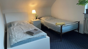 Une petite chambre à coucher, bien rangée, avec deux lits simples, une table de chevet avec une lampe, une plante en pot et un radiateur.