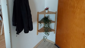 Un coin de pièce avec un porte-manteau sur lequel est accroché un manteau noir, une petite étagère en bois avec une plante en pot et une porte en bois.