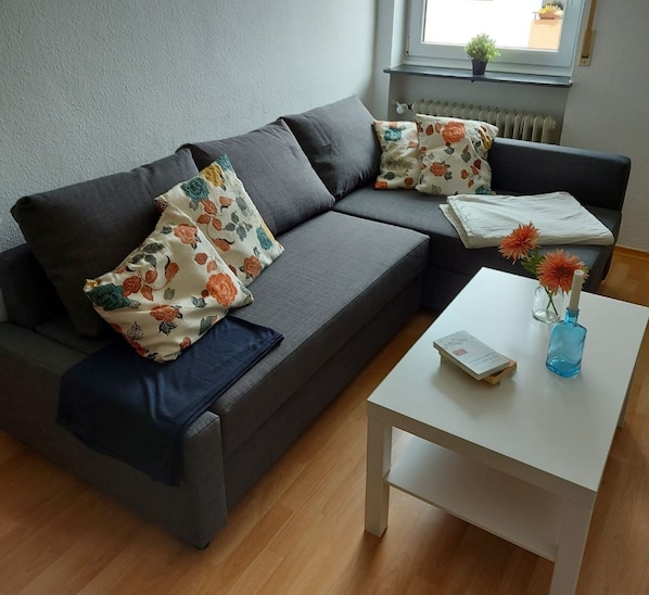 Een moderne woonkamer met een grijze bank, bloemenkussens, een witte salontafel en een kleine plant in een pot.