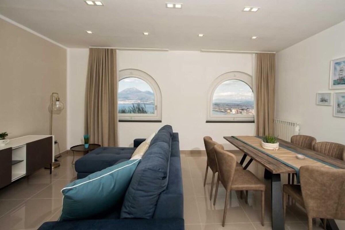Un salon moderne avec un canapé, une table à manger et des chaises. Il y a deux grandes fenêtres avec des rideaux, un radiateur et une plante sur la table.