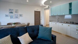 Een moderne woonkamer met een bank, een eettafel en een keukenhoek met blauwgroene kastjes en een spoelbak.