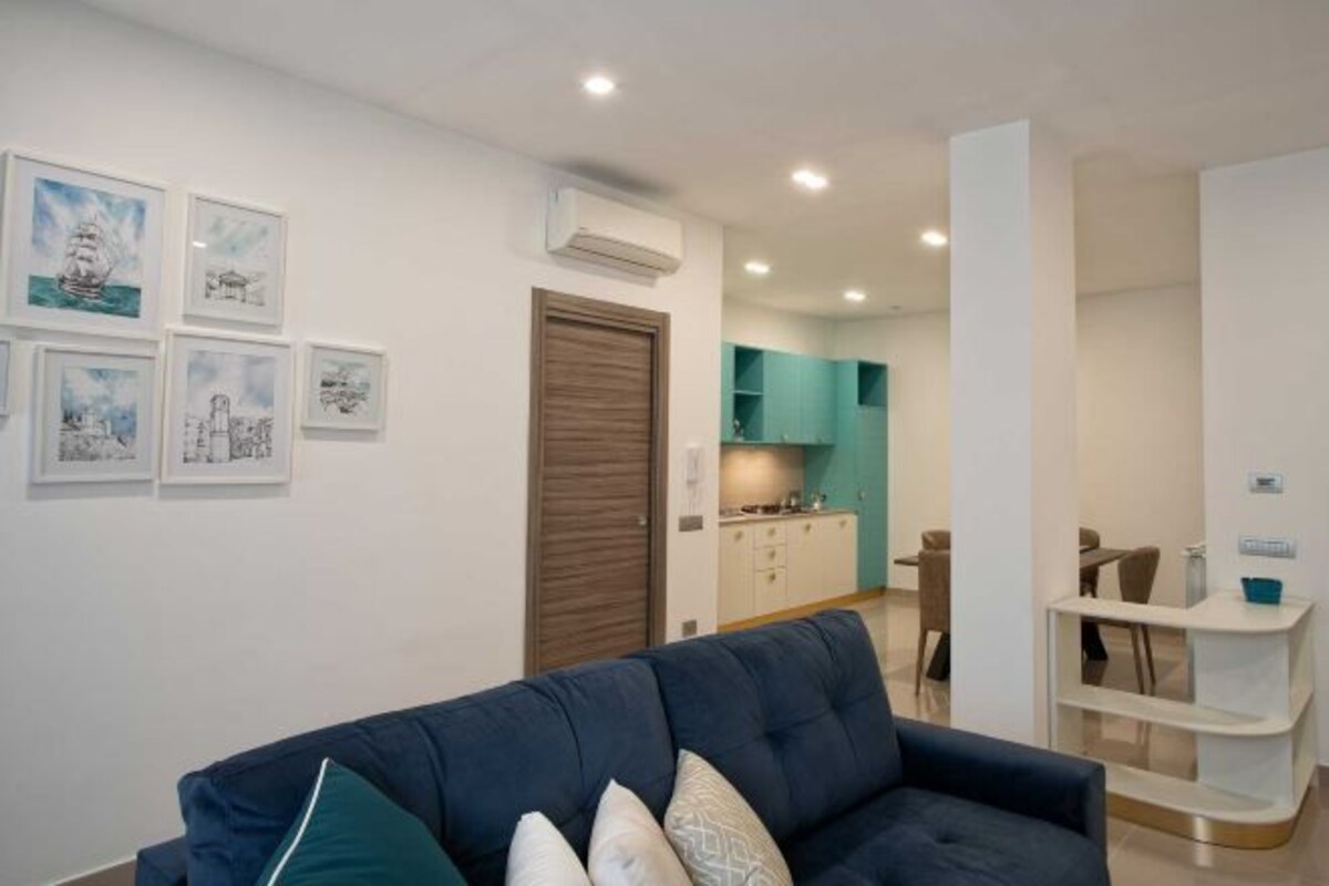 Un salon moderne avec un canapé bleu foncé, une porte en bois et un coin cuisine à l’arrière-plan.