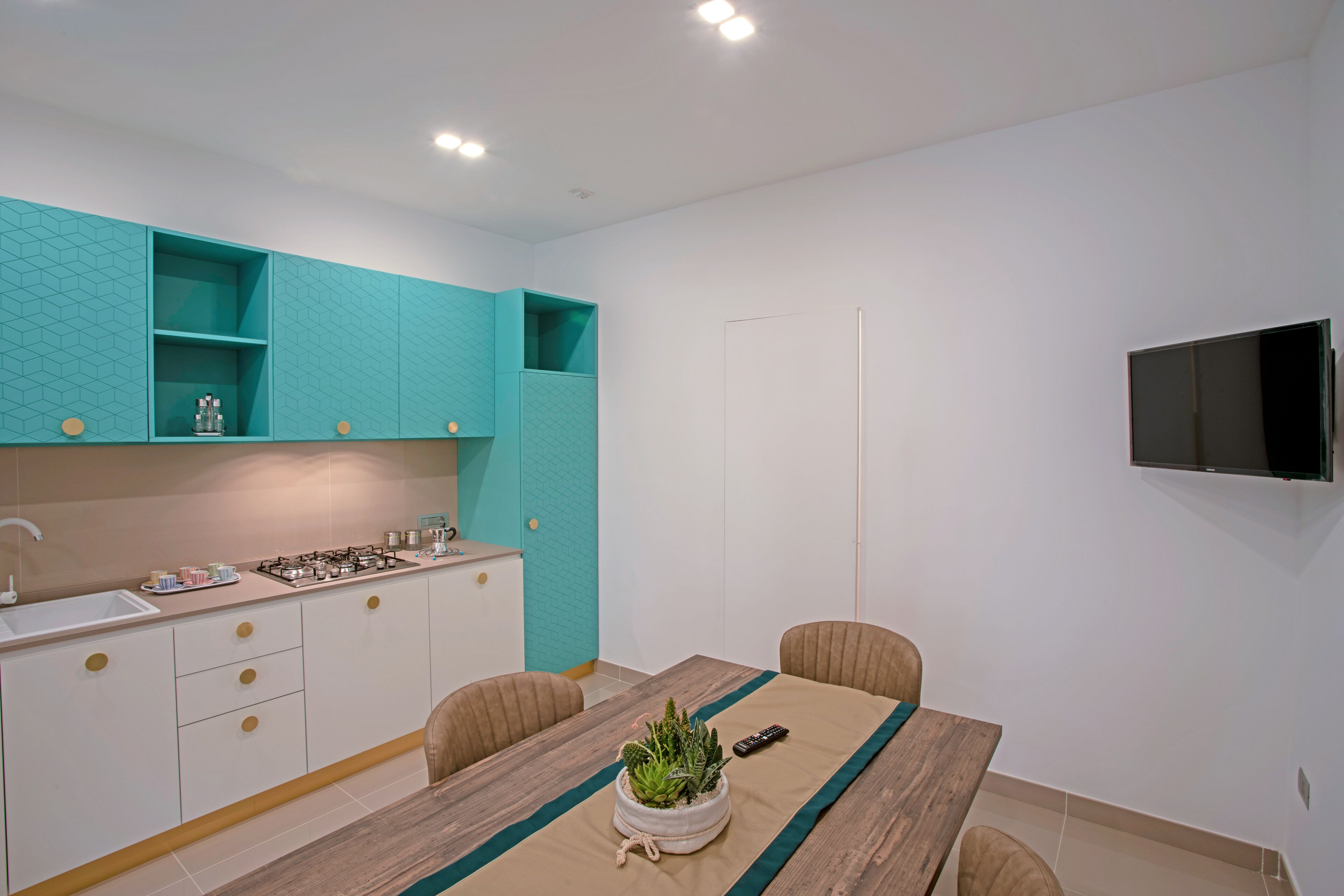 A modern kitchen with teal cabinetry, a wooden dining table with chairs, and a TV mounted on the wall.