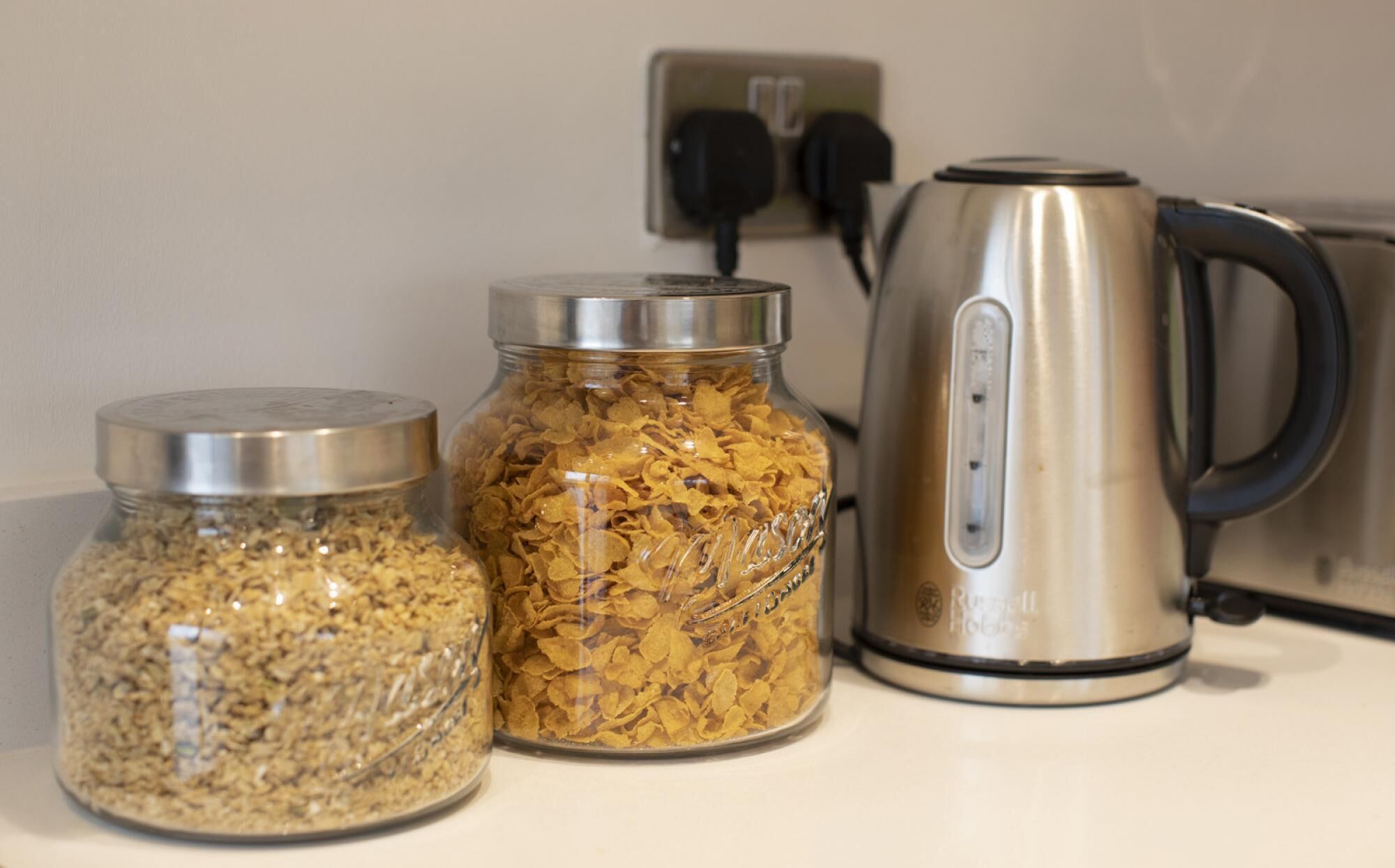 Two glass jars with metal lids containing different food items, a stainless steel electric kettle, and a power outlet with two plugs.