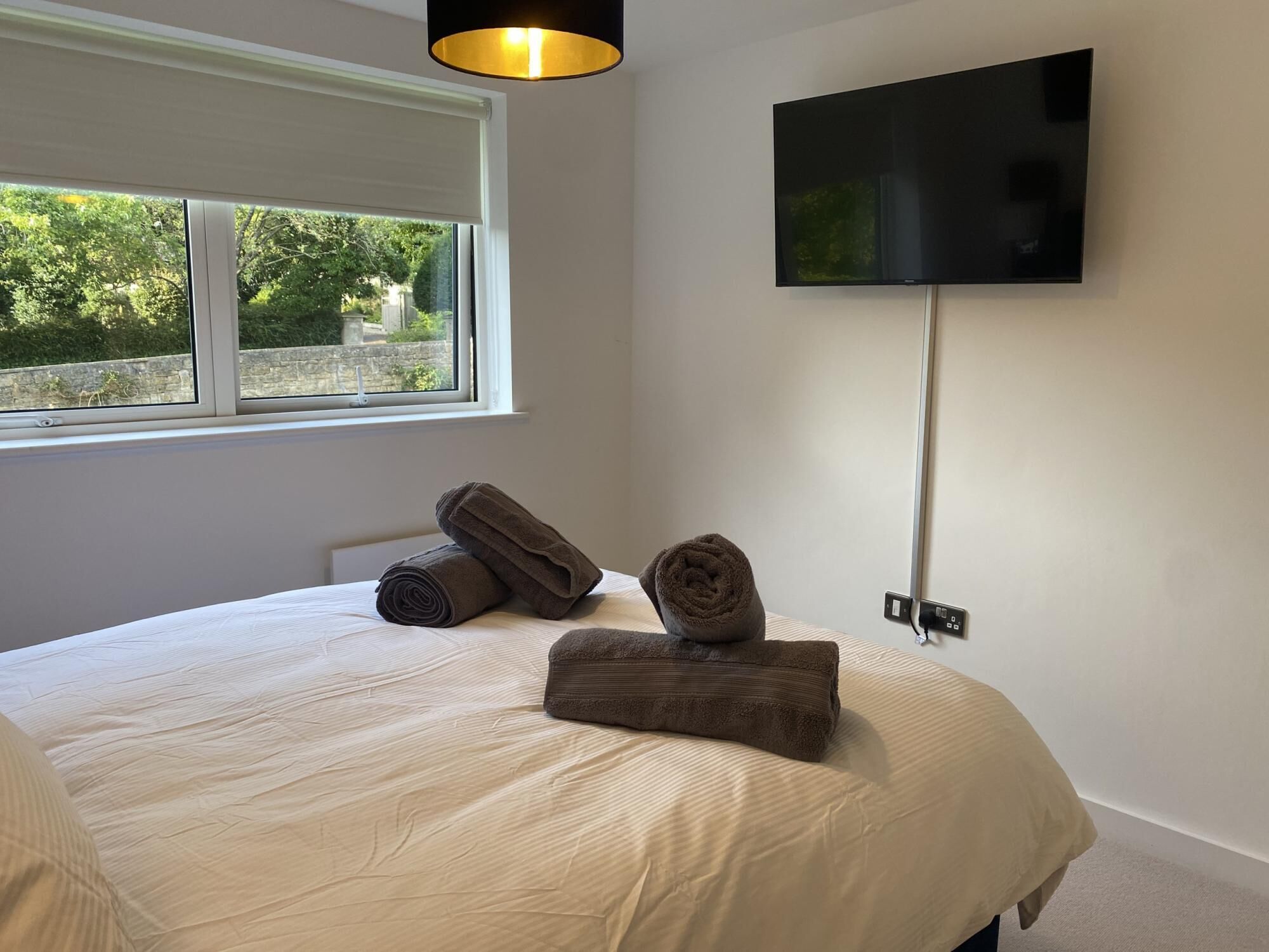 A bedroom with a bed, two rolled towels, a TV mounted on the wall, and a window with a view of greenery.
