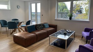 Un salon moderne avec un canapé en cuir marron, une table basse en verre et des fauteuils violets. On y trouve également un coin repas avec une table en verre et des chaises bleues. Une grande fenêtre offre une vue sur l’extérieur.