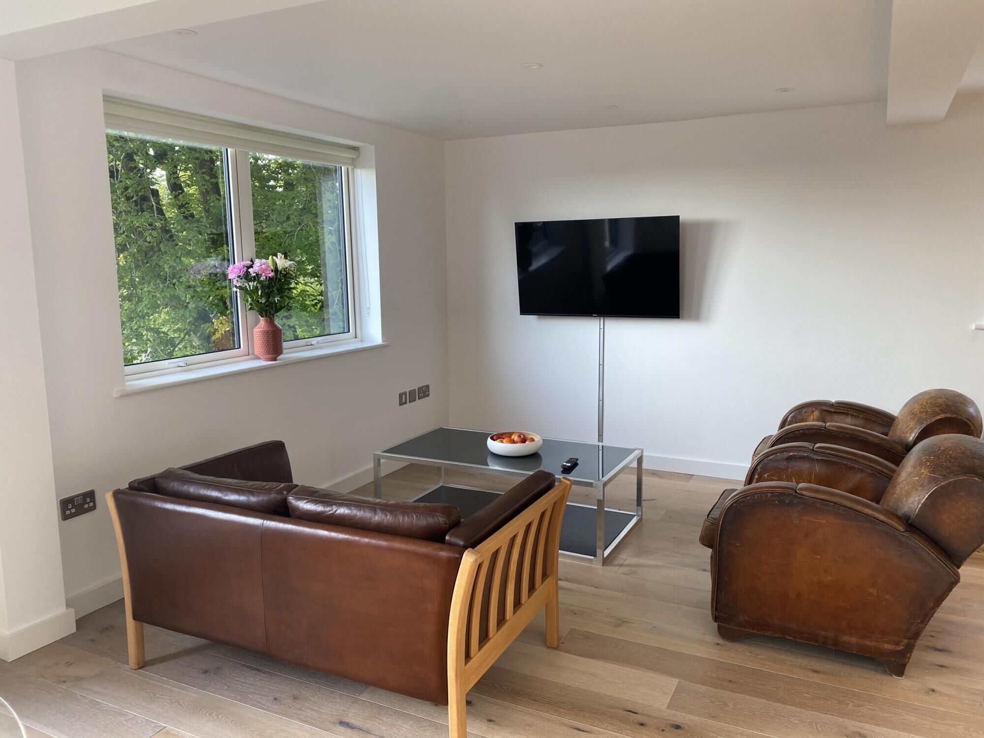 A modern living room with a leather sofa, a glass coffee table, a TV mounted on the wall, and a window with a view of greenery.