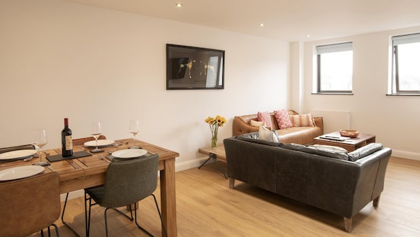 Een moderne woonkamer met een houten eettafel voor twee personen, een leren bank en een groot raam dat voor natuurlijk licht zorgt.
