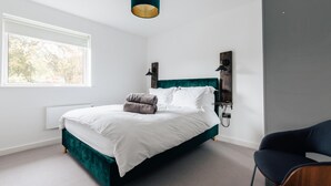 Un dormitorio moderno con una cama grande, cabecera verde oscuro, mesita de noche con lámpara y vistas a árboles a través de una ventana.