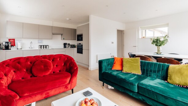 Una sala moderna con un sofá rojo y aguamarina, una mesa de centro blanca y una cocina al fondo.