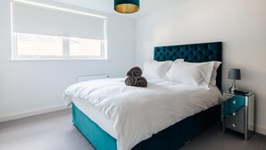 A modern bedroom with a teal upholstered bed, white bedding, a dark teal headboard, a bedside table with a lamp, and a window with blinds. - CH Apt 4 2bed in Bath (Bath)