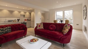 Un salon moderne avec deux canapés en velours rouge, une table basse blanche sur laquelle se trouve un saladier rempli de fruits, et une grande fenêtre donnant sur l’extérieur.