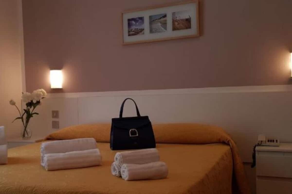Cama arrumada com toalhas e uma bolsa de mão preta, um vaso com flores brancas e um quadro na parede.