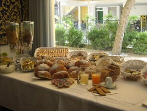 A table with various pastries and drinks, a window with a view of greenery and a building, and a tree outside. - Hotel Haiti B&B (Cervia)