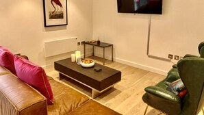 A modern living room with a flat-screen TV mounted on the wall, a brown leather sofa, a green leather armchair, a dark coffee table with candles, and a framed picture on the wall. - CH Apt 5 2bed in Bath (Bath)