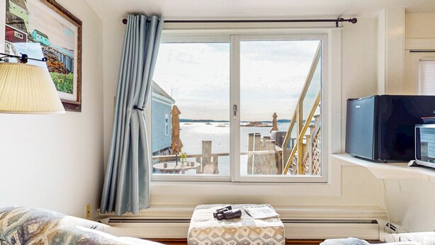 A room with a large window overlooking a deck with outdoor furniture, a television on a stand, and a table with a newspaper. - The Grace Bailey - Inn on the Harbor (Stonington)
