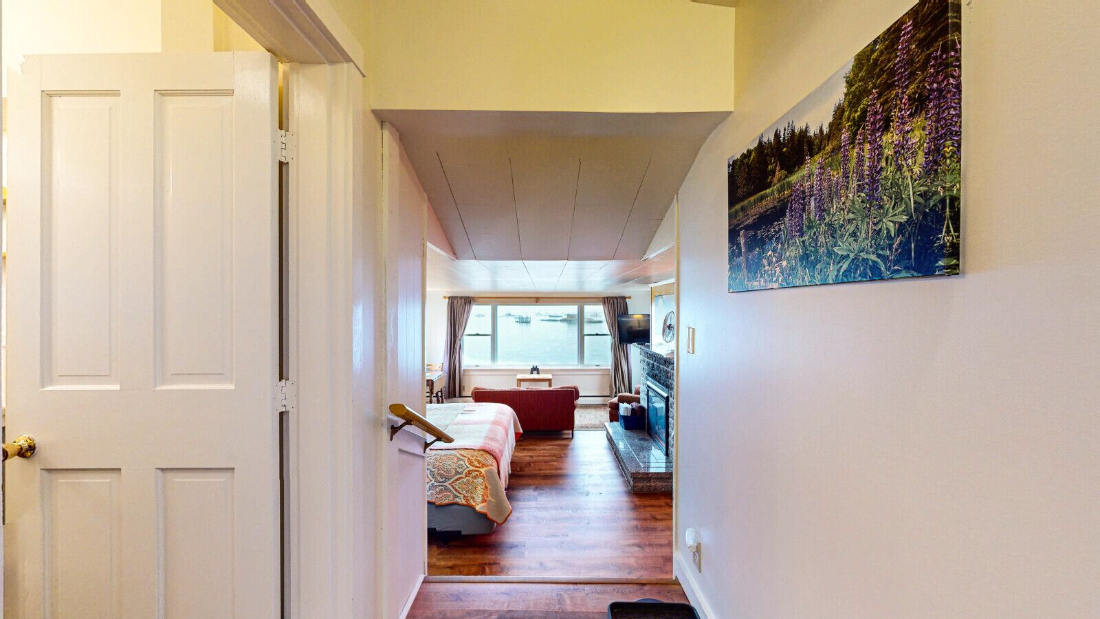 Un couloir étroit mène à une chambre dotée d’une grande fenêtre, d’un lit, d’un fauteuil rouge et d’un tableau accroché au mur.
