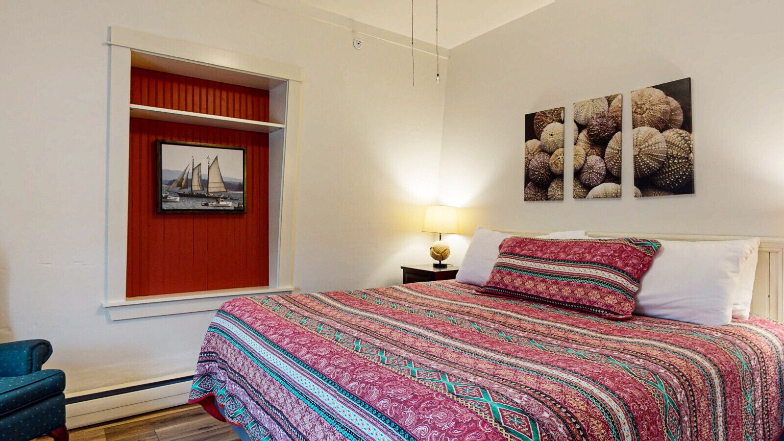 Ein Schlafzimmer mit einem Bett, einem Fenster mit rotem Rahmen, einem Nachttisch mit Lampe und Wandbildern.