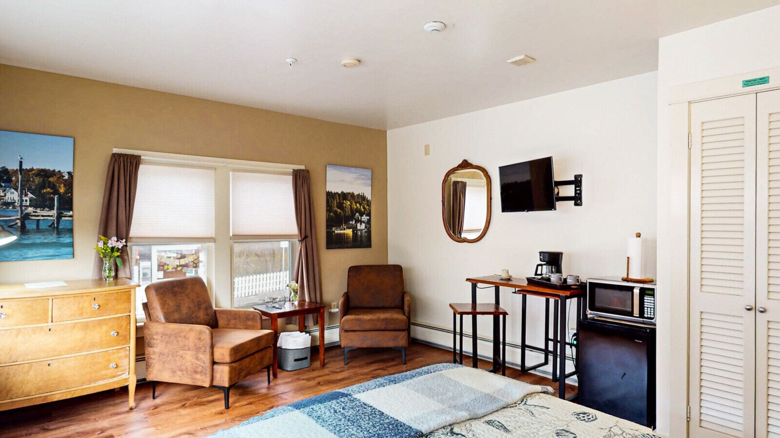 A cozy room with a wooden dresser, two brown armchairs, a small table with a coffee maker, a TV, and a window with curtains.