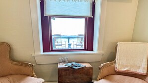 A room with two wicker chairs, a small wooden table with a magazine and glasses, and a window with a view of a balcony. - The Roseway - Inn on the Harbor (Stonington)