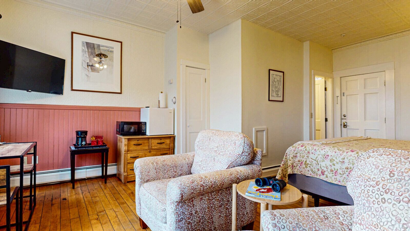Ein gemütliches Zimmer mit Holzboden, einer Couch, einem kleinen Tisch mit Buch und Kopfhörern, einer Mikrowelle und einem Fernseher an der Wand.