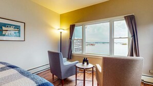Habitación de hotel con un ventanal grande, dos sillones, una mesita con unos prismáticos y un cuadro con un barco en la pared.