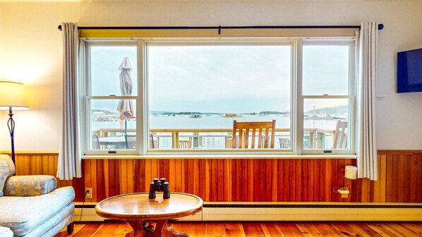 A room with a large window overlooking a waterfront, a round wooden table with binoculars, a chair, and a lamp.