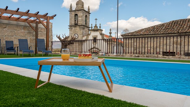 A poolside breakfast setup with a table, a glass of orange juice, and a plate of food, overlooking a historic church and a stone building. - The Village House by CDV (Carrazeda de Ansiaes)