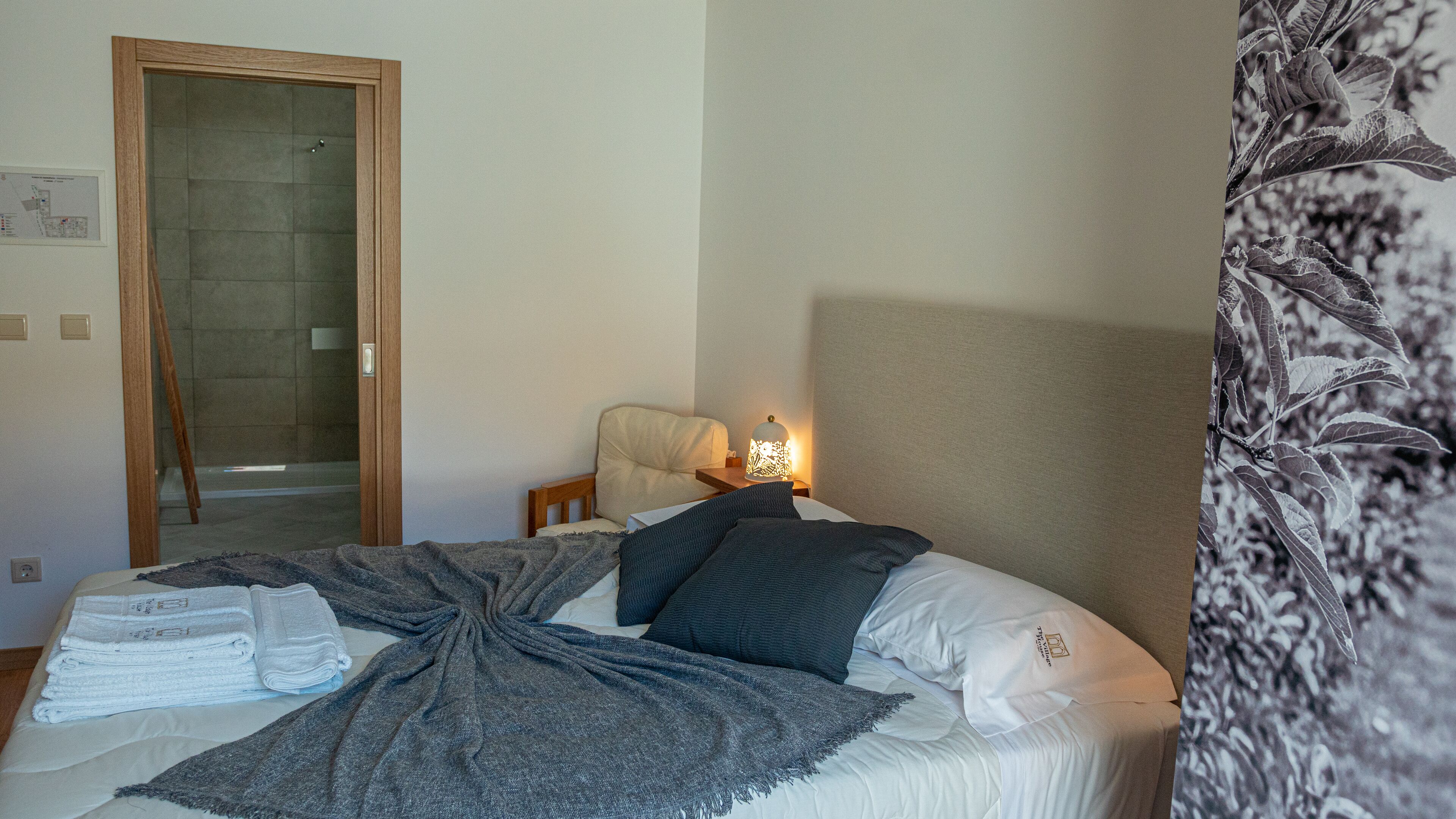 Una habitación de hotel con una cama, una silla, una mesita de noche con lámpara y una puerta que da a una zona de ducha.