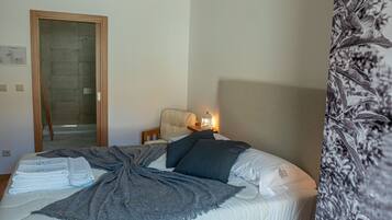 Una habitación de hotel con una cama, una silla, una mesita de noche con lámpara y una puerta que da a una zona de ducha.