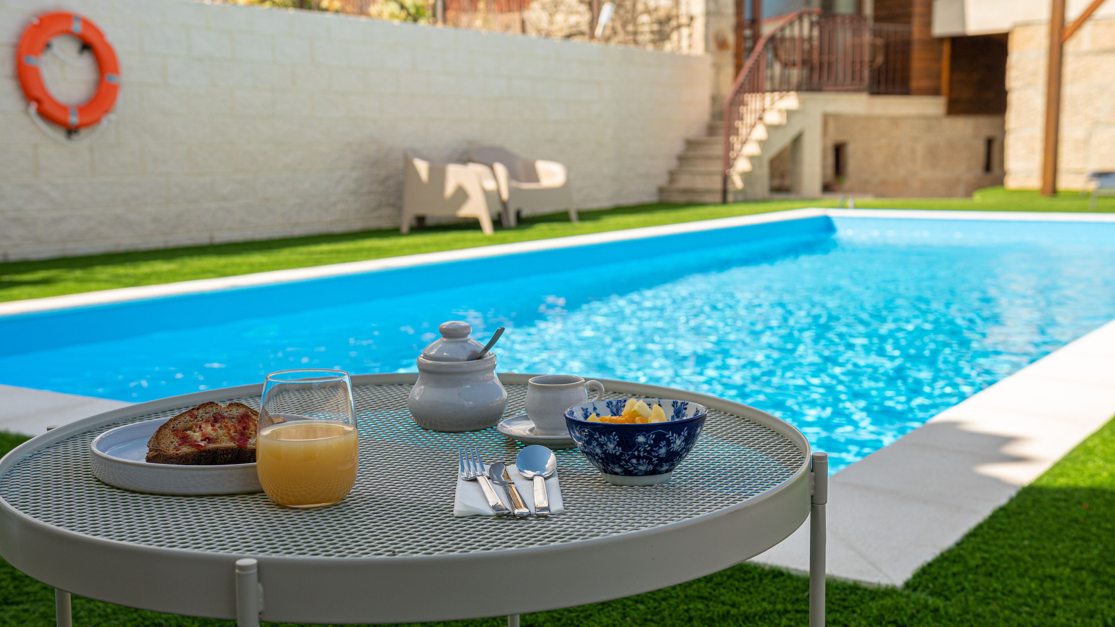 Mesa metálica redonda con alimentos para el desayuno, como tostadas, un vaso de jugo de naranja, una tetera, una taza y un tazón de fruta, junto a una piscina.