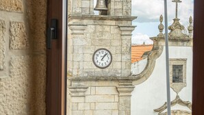 A window with a view of a historic clock tower, a wooden table with glasses and a remote control, and a stone wall. - The Village House by CDV (Carrazeda de Ansiaes)