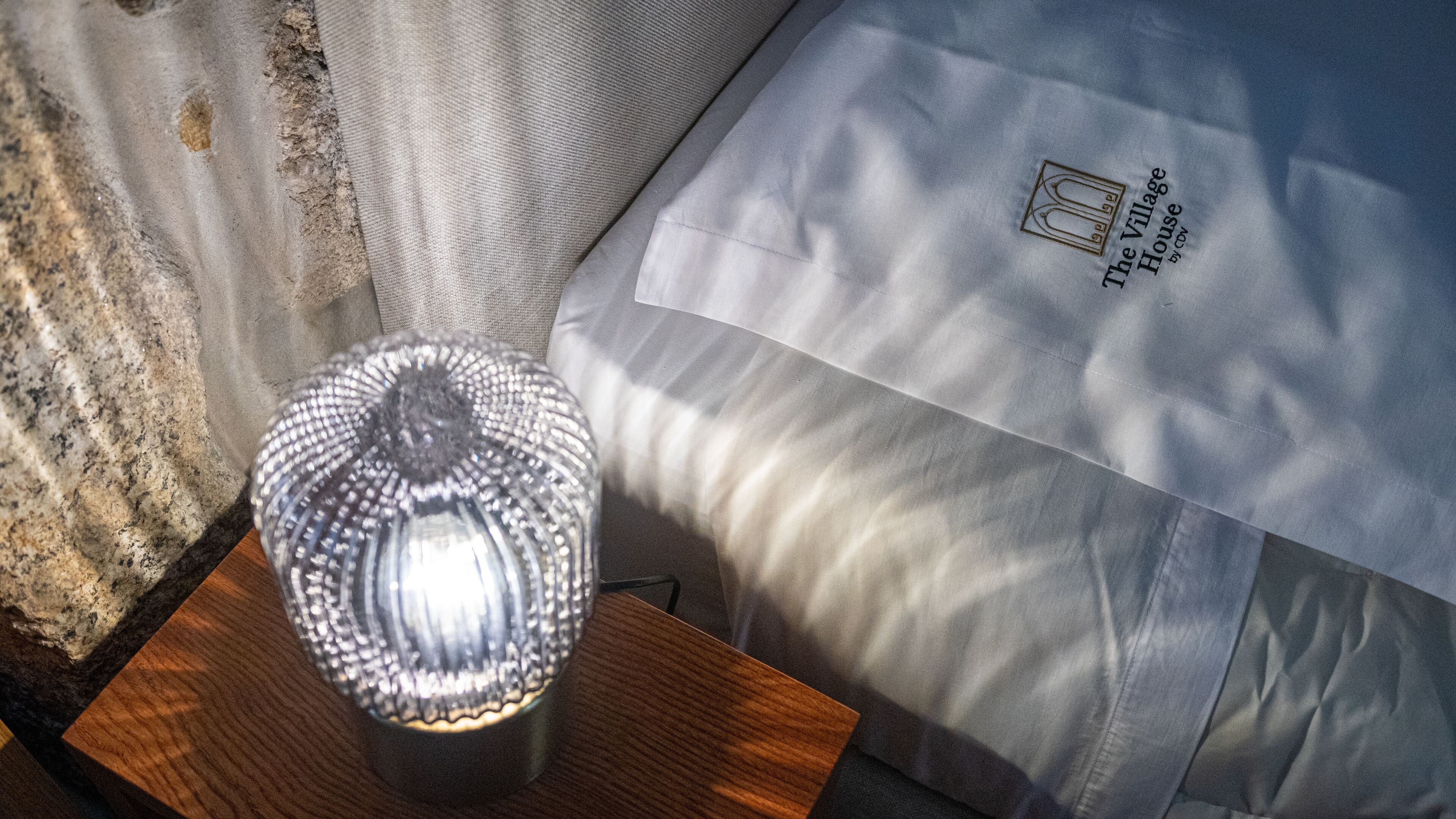 A lamp with a textured glass shade on a wooden surface, next to a bed with a pillow that has a logo and text.