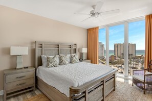 Une chambre dotée d’un grand lit, de deux tables de chevet, d’un ventilateur de plafond et offrant une vue sur la ville et la plage.