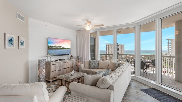 Ein großzügiges Wohnzimmer mit einem großen Flachbildfernseher, einer Couch und einem Couchtisch. Große Fenster bieten Blick auf das Meer und die Stadt.