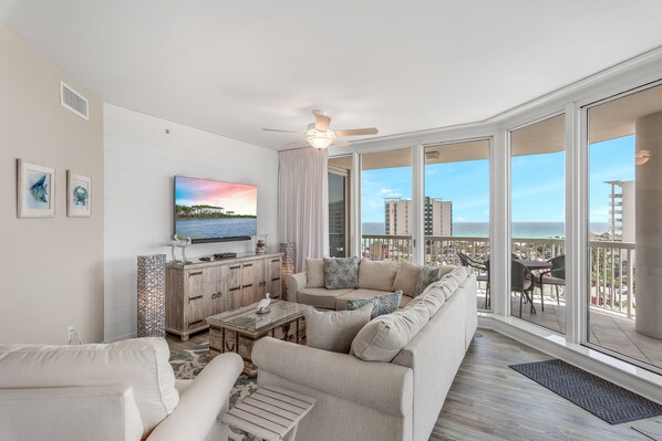A spacious living room with a large flat-screen TV, a sofa, and a coffee table. There are large windows offering a view of the ocean and buildings.