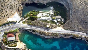 An aerial view of a coastal area with a large natural sinkhole, a bridge, and a building complex with parking and recreational areas. - Seaside Nestie by BluPine (Vari-Voula-Vouliagmeni)