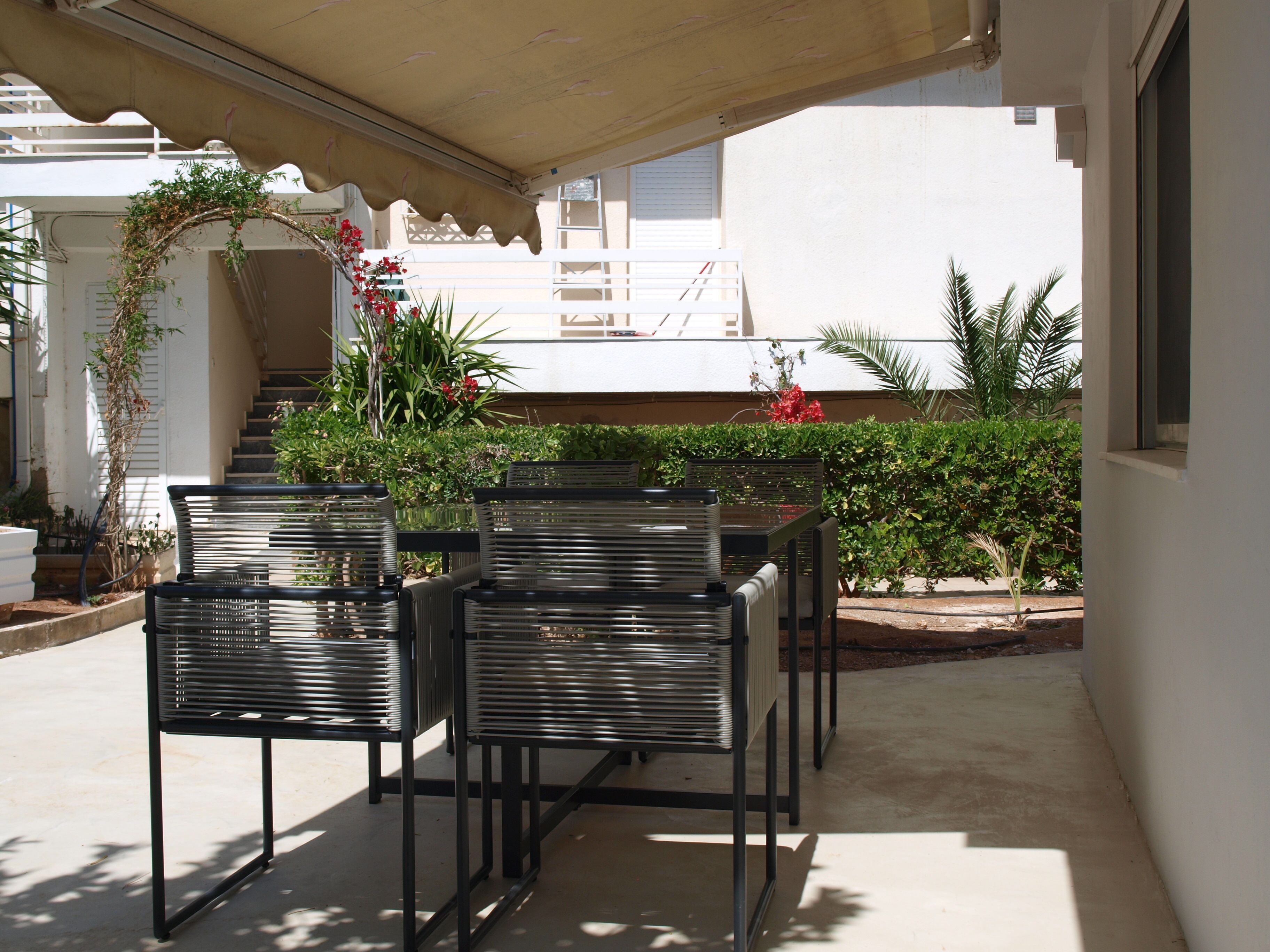 A covered outdoor seating area with metal chairs and tables, surrounded by greenery and a staircase leading to an upper level.
