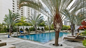 Zona de piscina con sillones de descanso, palmeras y una piscina azul cristalina rodeada de una terraza con baldosas.