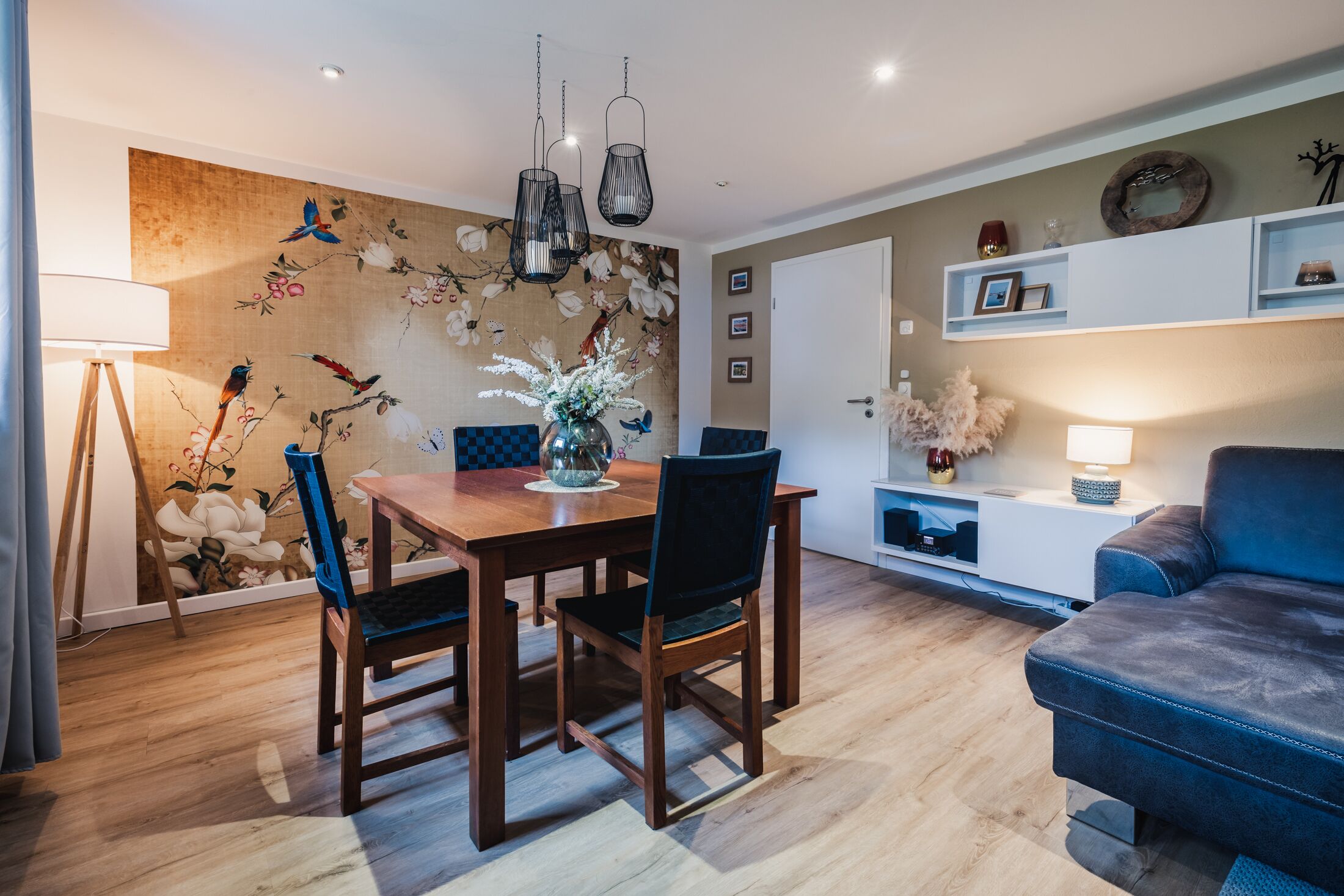 A modern living room with a wooden dining table, two chairs, a blue sofa, and a decorative wall with a floral mural.