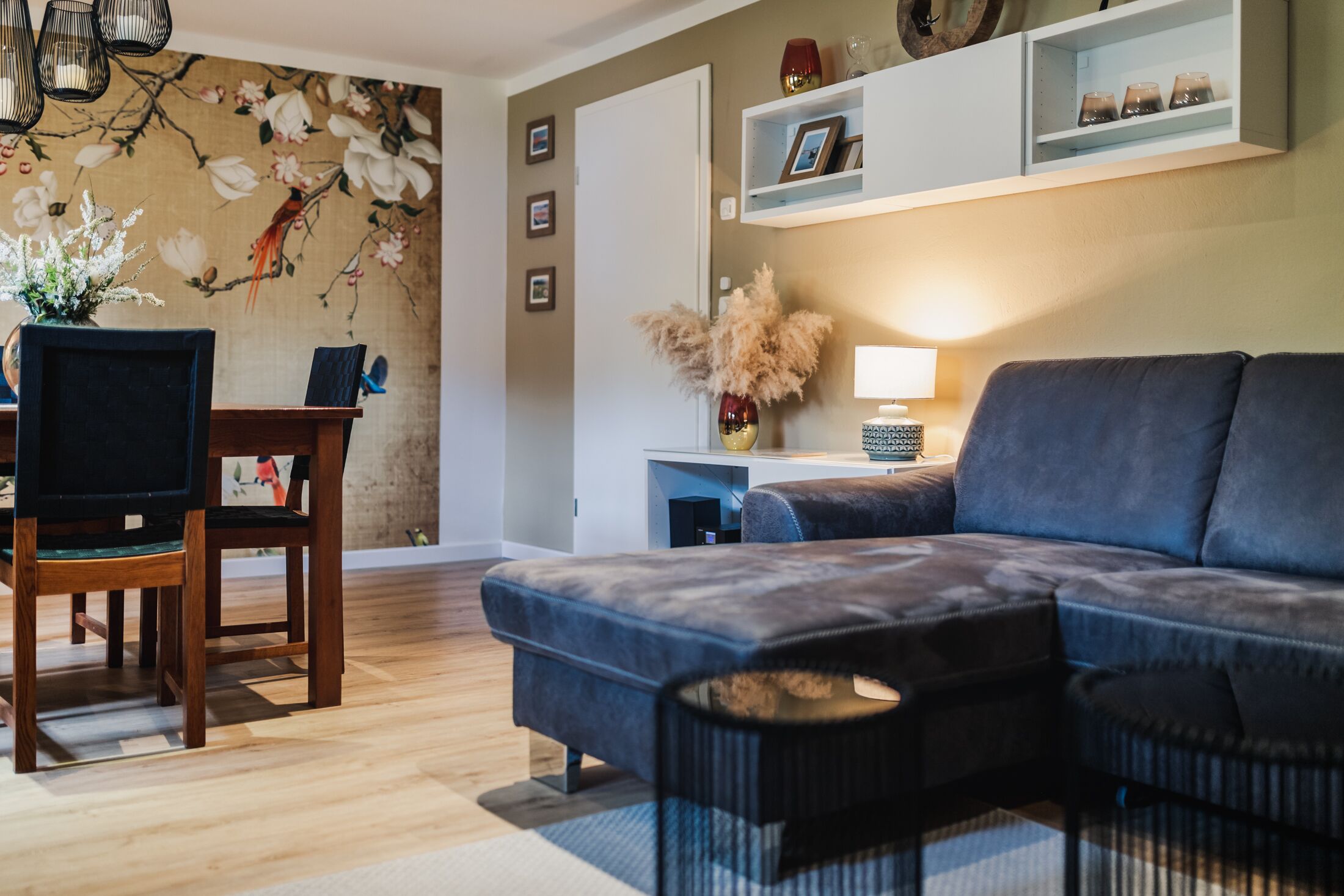 A modern living room with a sectional sofa, a wooden dining table, and a wall-mounted shelf with decorative items.
