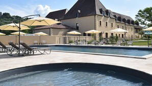 A swimming pool area with lounge chairs and umbrellas, a large building with a sloped roof, and a clear sky. - Hotel le Périgord (La Roque-Gageac)