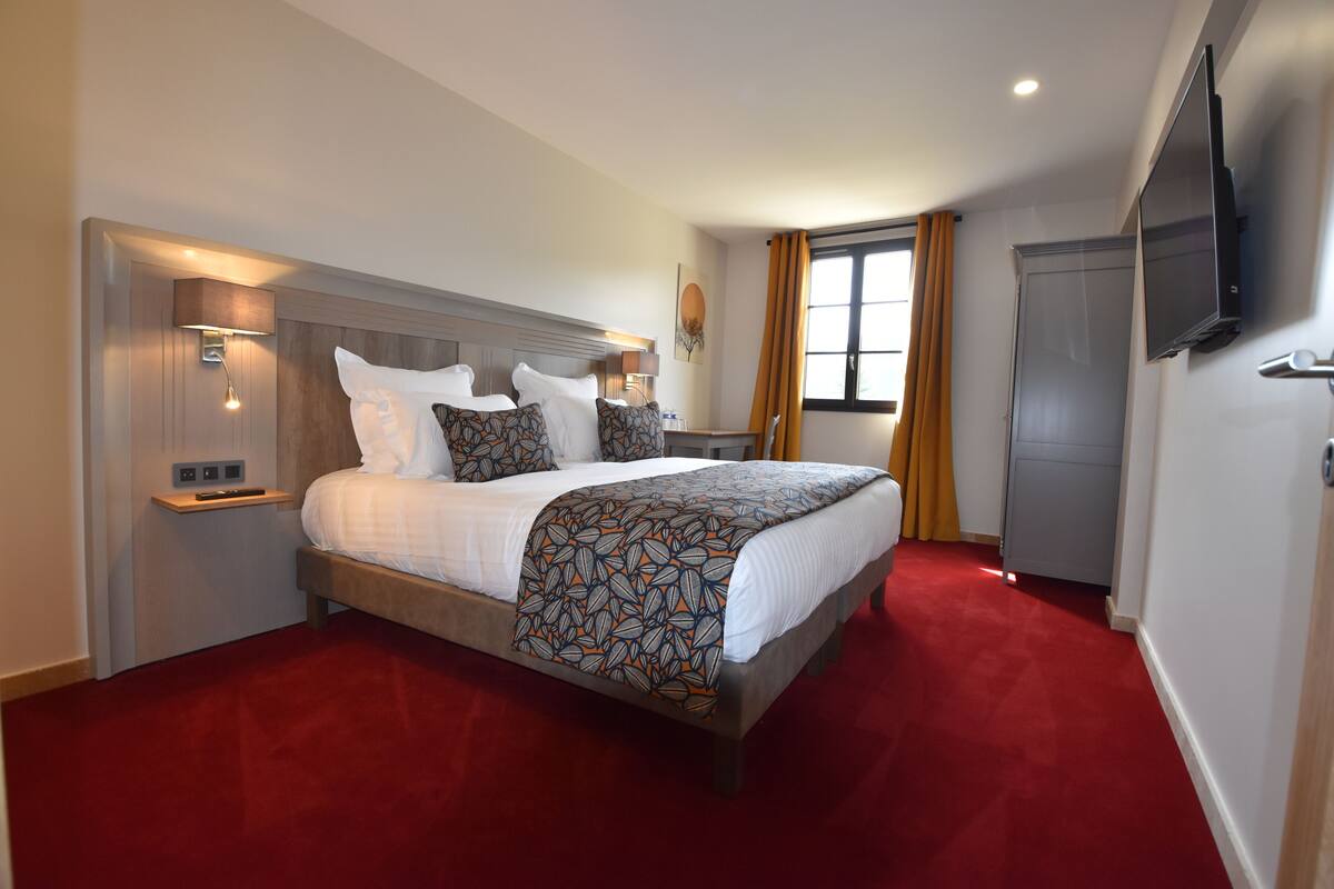 A hotel room with a large bed, a TV mounted on the wall, a desk with a lamp, and a window with curtains.
