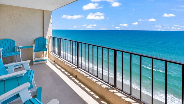 Un balcon avec des chaises turquoise donnant sur une plage aux eaux bleu clair, le tout agrémenté d’une rambarde en métal.